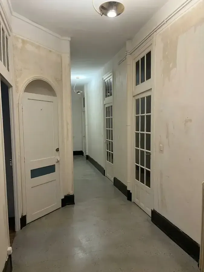 Couloir ancien avant travaux : murs tachés et cloqués, sol en béton usé, portes vitrées et corniches encrassées