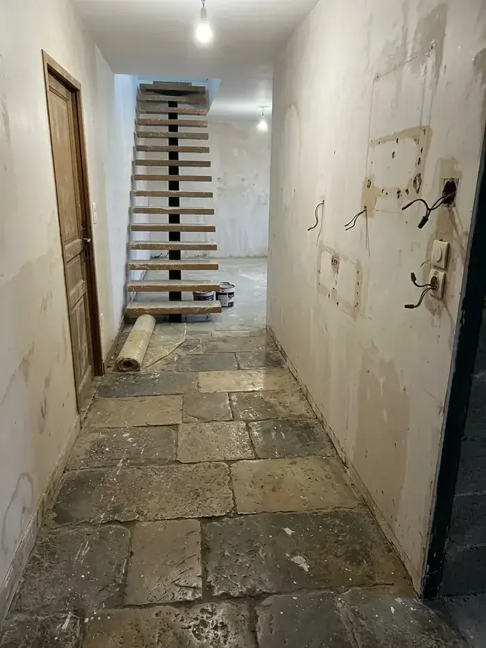 Entrée en travaux : murs décroûtés, boîtiers électriques et fils sortis, dalles de pierre disjointes, escalier nu