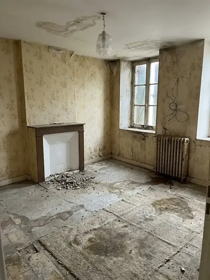 Pièce très dégradée : papier peint fleuri décollé, auréoles d'infiltration au plafond, sol éventré, radiateur en fonte rouillé