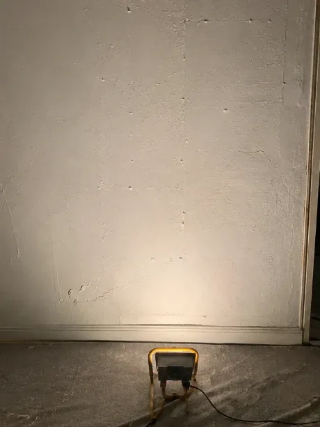 Mur éclairé par un projecteur posé au sol, avant préparation : joints, rebouchages et coups de couteau ressortent en ombres